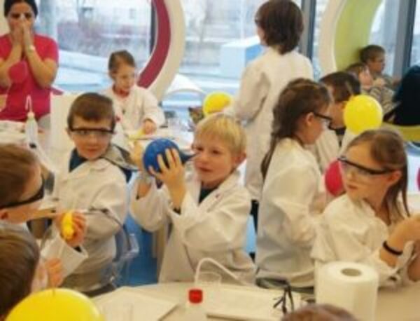 Kinder in Laborkitteln und mit Schutzbrillen nehmen an einer wissenschaftlichen Aktivität an einem Tisch im Klassenzimmer teil und hantieren mit Luftballons und verschiedenen Laborgeräten.