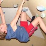 Ein Kind in blauem Hemd und roten Shorts klettert an einer Indoor-Felswand und hält sich mit beiden Händen und Füßen fest.
