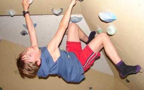 Ein Kind in blauem Hemd und roten Shorts klettert an einer Indoor-Felswand und hält sich mit beiden Händen und Füßen fest.