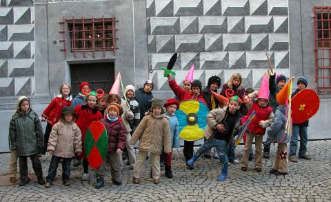 Eine Gruppe von Kindern posiert im Freien in Winterkleidung, mit bunten Pappschilden und Schwertern in der Hand, einige tragen Kegelhüte, vor einer gemusterten Wand.