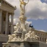 Die Athena-Statue steht auf einem Brunnen vor dem österreichischen Parlamentsgebäude, mit neoklassizistischer Architektur und bewölktem Himmel im Hintergrund.