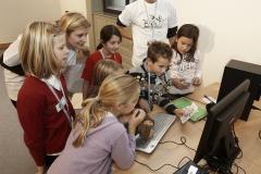 Eine Gruppe von Kindern versammelt sich unter Aufsicht eines Erwachsenen um einen Computermonitor, während sie an einem Schreibtisch eine Tätigkeit ausüben.