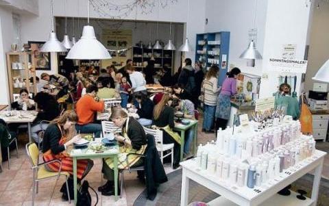 Ein belebtes Café mit Menschen, die an Tischen sitzen, essen und sich unterhalten; Regale mit Produkten sind sichtbar, und helle Oberlichter erhellen den Raum.