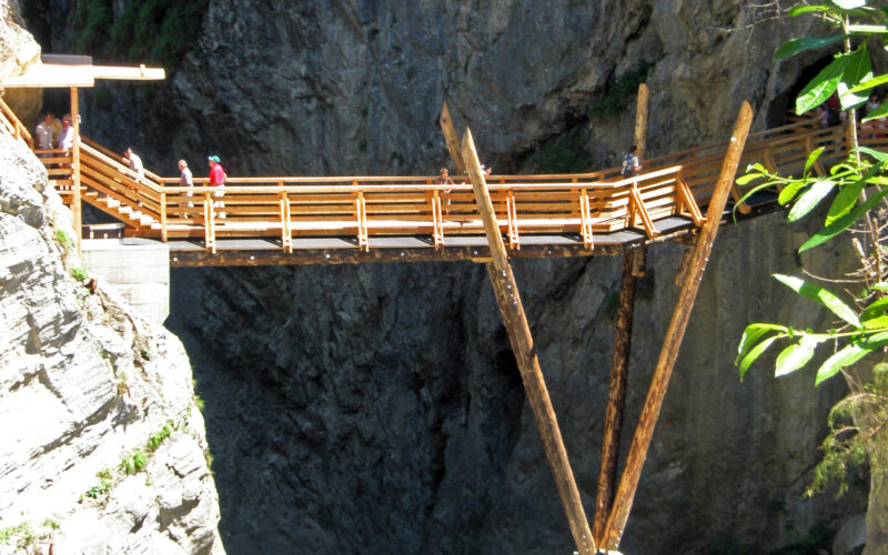 Eine hölzerne Fußgängerbrücke mit abgewinkelten Stützbalken überspannt eine felsige Schlucht über einem fließenden Fluss, über die Menschen gehen.