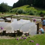 Zwei Kinder spielen in der Nähe eines flachen Teichs im Freien, umgeben von hölzernen Trittsteinen, einem Seilnetz und einer Graslandschaft mit Bäumen im Hintergrund.