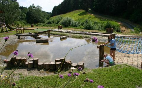 Zwei Kinder spielen in der Nähe eines flachen Teichs im Freien, umgeben von hölzernen Trittsteinen, einem Seilnetz und einer Graslandschaft mit Bäumen im Hintergrund.