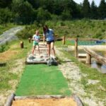 Zwei Personen spielen an einem sonnigen Tag im Freien Minigolf, umgeben von Grün und Bäumen.