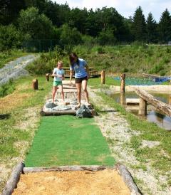 Zwei Personen spielen an einem sonnigen Tag im Freien Minigolf, umgeben von Grün und Bäumen.