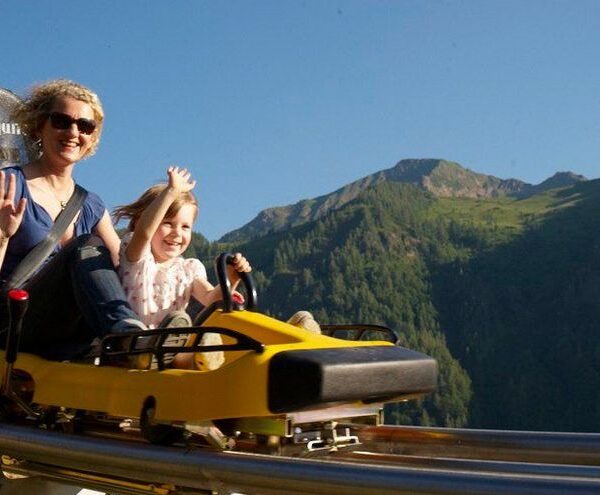 Zwei Personen, ein Erwachsener und ein Kind, fahren lächelnd und winkend mit Bergen und Bäumen im Hintergrund auf einer gelben Alpen-Achterbahn im Freien.