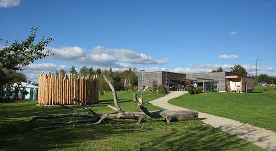 Ein modernes Gebäude mit einer Holzfassade steht auf einer Wiese, neben einem umgestürzten Baumstamm und einem runden Holzzaun, unter einem blauen Himmel mit vereinzelten Wolken.