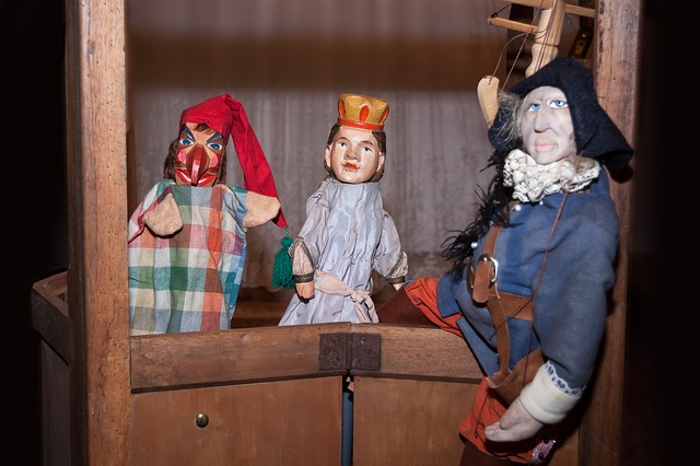 Drei Handpuppen sind auf einer kleinen hölzernen Puppentheaterbühne angeordnet, darunter eine Figur mit rotem Hut, eine Königin mit Krone und eine Figur in blauem Gewand mit Federhut.