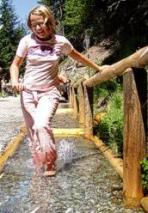 Eine Person läuft barfuß durch einen seichten Wasserlauf auf einem Steinweg und hält sich dabei an einem Holzgeländer im Freien fest.