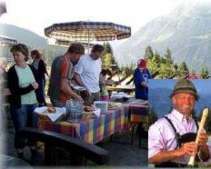 An einem Buffet im Freien in den Bergen werden Speisen serviert. Eine Einblendung zeigt einen Mann in traditioneller Alpenkleidung, der eine Holzflöte spielt.