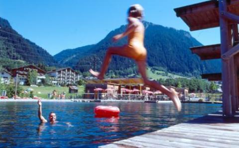 Ein Kind im Badeanzug springt von einem Steg in einen See, während eine Person vom Wasser aus winkt, mit Bergen und Gebäuden im Hintergrund.