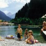 Kinder spielen am Rande eines Sees, der von Bergen umgeben ist, während die Erwachsenen auf einer Holzterrasse in der Nähe unter einem sonnigen Himmel sitzen.