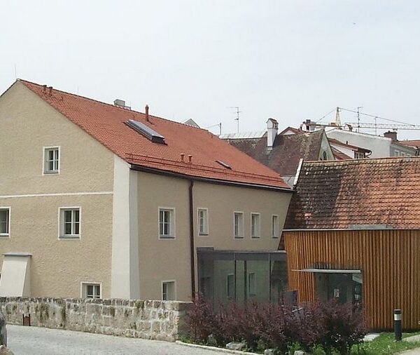 Eine Gruppe von Gebäuden mit roten Ziegeldächern, darunter ein beigefarbenes verputztes Haus und ein holzgetäfeltes Gebäude neben einem hohen, weißen rechteckigen Turm.
