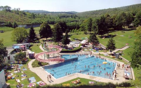 Freibad mit Menschen, die schwimmen und sich sonnen, umgeben von Rasenflächen, Bäumen und Hügeln im Hintergrund an einem sonnigen Tag.
