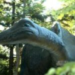 Eine große, realistische Dinosaurier-Statue steht inmitten von grünen Bäumen und Pflanzen im Zoo HAPP, wobei das Sonnenlicht durch die Blätter fällt.