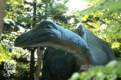 Eine große, realistische Dinosaurier-Statue steht inmitten von grünen Bäumen und Pflanzen im Zoo HAPP, wobei das Sonnenlicht durch die Blätter fällt.