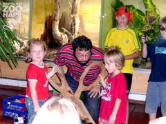 Ein Erwachsener kniet neben Kindern, die ein Rentiergeweih im Haus berühren, während im Hintergrund eine Zoo-Kulisse zu sehen ist.
