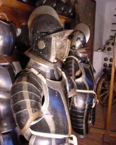 Zwei mittelalterliche Rüstungen mit Helmen und Visieren sind in einem Museum nebeneinander ausgestellt, umgeben von anderen historischen Rüstungsteilen.