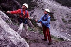 Zwei Personen mit Helm und Klettergurt stehen in felsigem Gelände; einer steigt die Felswand hinauf, während der andere das Seil zur Sicherung hält.
