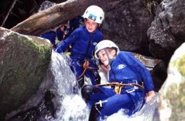 Zwei Personen, die Helme und Neoprenanzüge tragen, navigieren in einem felsigen Bach, umgeben von Wasser und großen Felsbrocken in einer Außenumgebung.