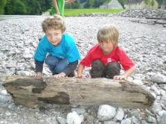 Zwei kleine Kinder kauern auf einer felsigen Fläche im Freien und stützen sich jeweils mit einer Hand auf ein großes Stück Treibholz.