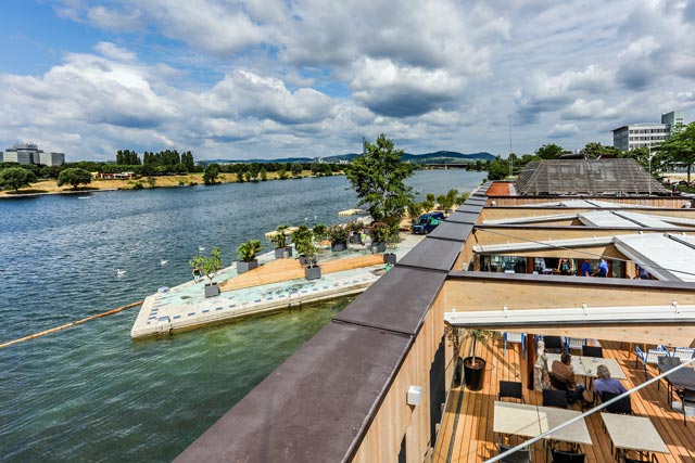 Eine Szene am Flussufer mit einem Steg, Sitzgelegenheiten und Menschen, die sich am Wasser unter einem teilweise bewölkten Himmel entspannen. Gebäude und Bäume säumen das gegenüberliegende Flussufer.