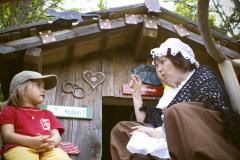 Eine ältere Frau in einem Kostüm spricht mit einem Kind, das vor einem kleinen Holzhaus sitzt, das mit Herz- und Brillenschildern geschmückt ist.