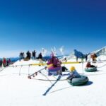 Menschen vergnügen sich beim Snowtubing und anderen Winteraktivitäten auf einem verschneiten Berghang unter strahlend blauem Himmel, mit weit entfernten Gipfeln im Hintergrund.