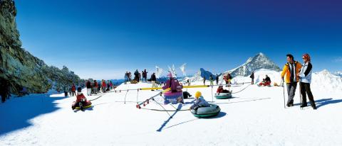 Menschen vergnügen sich beim Snowtubing und anderen Winteraktivitäten auf einem verschneiten Berghang unter strahlend blauem Himmel, mit weit entfernten Gipfeln im Hintergrund.
