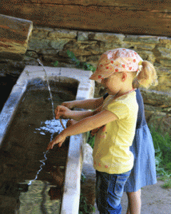 Zwei kleine Kinder stehen nebeneinander und spielen mit dem Wasser, das aus einem Ausguss in ein Steinbecken im Freien fließt.