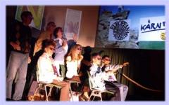 Sieben Personen, teils sitzend, teils stehend, stehen mit Sonnenbrillen auf der Bühne, hinter ihnen sind projizierte Bilder und Zeichen zu sehen.