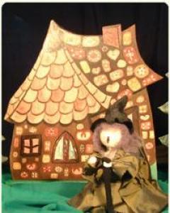 Eine als Hexe verkleidete Puppe steht vor einer Kulisse, die ein mit Süßigkeiten und Keksen dekoriertes Lebkuchenhaus zeigt.