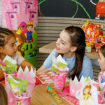 Drei Mädchen sitzen an einem dekorierten Tisch mit rosa Prinzessinnen-Partyzubehör und unterhalten sich während einer Geburtstagsfeier mit eingepackten Geschenken im Hintergrund.
