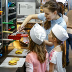 Eine Frau weist zwei Kinder mit Kochmützen an, wie man Burger an der Theke eines Schnellrestaurants zusammenstellt.