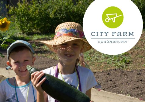 Zwei Kinder mit Hüten halten eine große Zucchini in einem Garten, in der oberen rechten Ecke ist das Logo der City Farm Schönbrunn zu sehen.