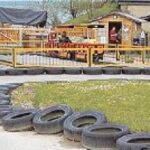 Kinder fahren kleine Gokarts auf einer von Reifen gesäumten Strecke auf einem Vergnügungsgelände mit verschiedenen Attraktionen und Menschen im Hintergrund.