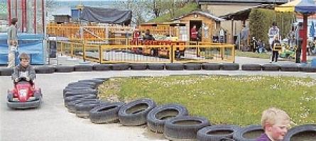 Kinder fahren kleine Gokarts auf einer von Reifen gesäumten Strecke auf einem Vergnügungsgelände mit verschiedenen Attraktionen und Menschen im Hintergrund.