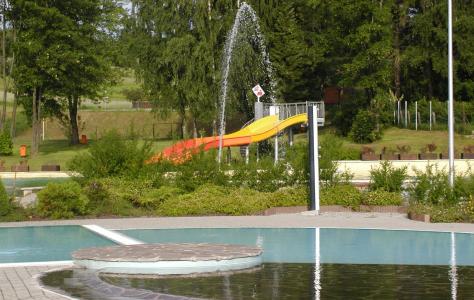 Freibad mit kreisförmigen Plattformen, einem Springbrunnen, der Wasser versprüht, und einer gelb-orangenen Rutsche im Hintergrund, umgeben von Grün und Bäumen.