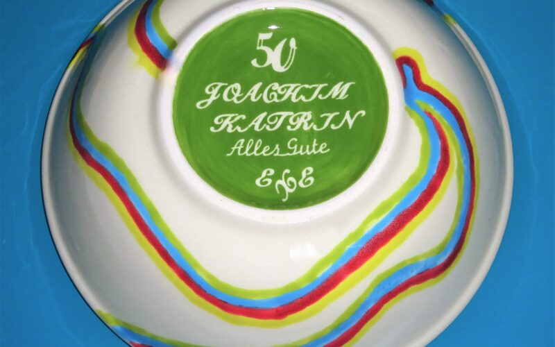 Eine weiße Schale mit bunten Wellenlinien, grünem Boden und weißer Schrift "50 Joachim Katrin Alles Gute 666" auf blauem Hintergrund.