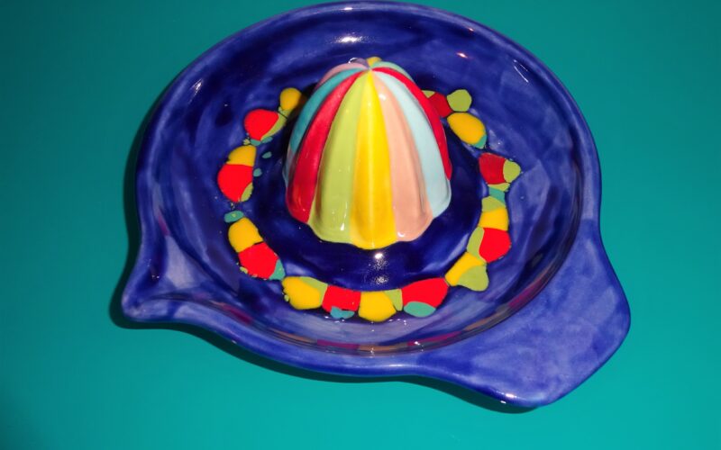 Eine farbenfrohe Zitruspresse aus Keramik steht auf einer tealfarbenen Oberfläche. Der Entsaftungskegel ist mit vertikalen Streifen in Rot, Gelb, Blau und Weiß versehen.