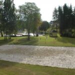 Im Freien ist ein Sandvolleyballfeld auf einer Wiese aufgebaut, umgeben von Bäumen und einigen Gebäuden im Hintergrund.