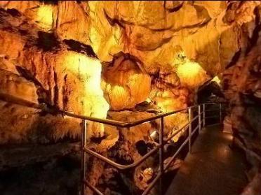 Ein gut beleuchteter Metallsteg führt durch das Innere einer Höhle mit orangefarbenen und braunen Felswänden.