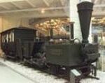 Schwarze Dampflokomotive und angehängter Personenwagen, die in einem Museum ausgestellt sind, mit einer weißen Decke und moderner Beleuchtung im Hintergrund.