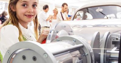 Ein junges Mädchen steht bei einer Indoor-Veranstaltung neben einer transparenten Maschine oder einem Exponat, während im Hintergrund Menschen und ein Auto zu sehen sind.