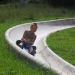 Ein kleines Kind sitzt auf einer von Gras umgebenen Betonrutsche und schaut nach vorne, während es sich an den Knien festhält.