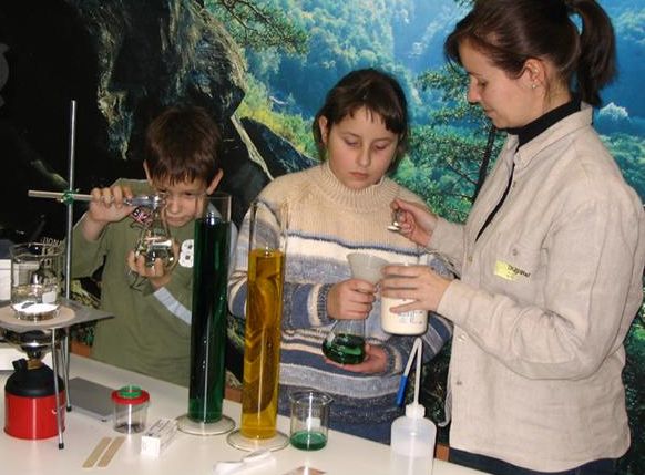 Eine Frau beaufsichtigt zwei Kinder, die an einem Labortisch wissenschaftliche Experimente mit Bechern und Messzylindern durchführen.