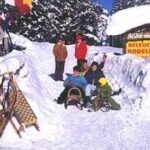 Eine Gruppe von Menschen mit Schlitten sitzt und steht an einem verschneiten Eingang, der mit einem gelben Schild "Beleuchtete Rodelbahn" gekennzeichnet ist, umgeben von Schnee und Bäumen.